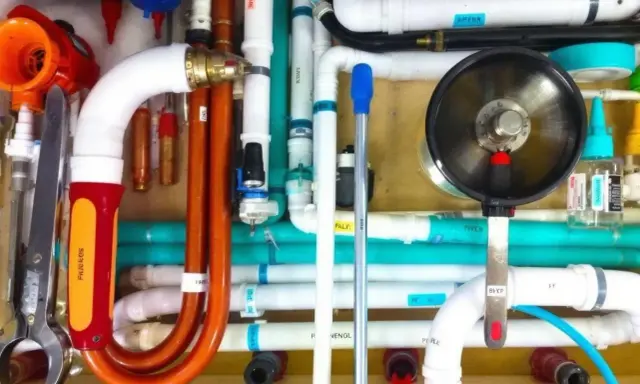 Un réseau complexe de tuyaux blancs, orange et turquoise, avec un outil de plomberie et un manomètre. Un **profilé souple** orange se courbe gracieusement.