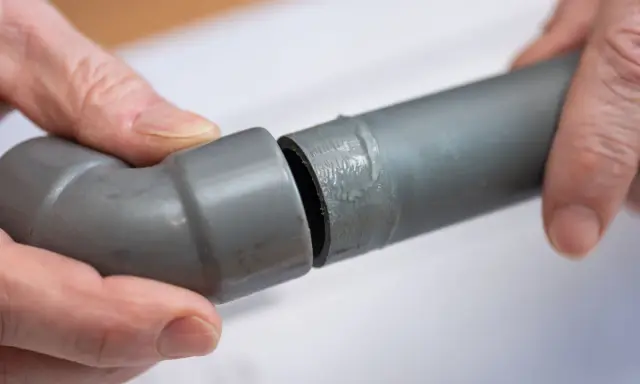 Mains de personne assemblant deux tuyaux gris en **pvc renforcé**, l'un avec un coude, prêts pour une installation solide.