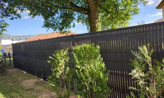 Clôture ajourée noire, offrant une protection discrète pour les tuyaux PVC. Des arbustes verts bordent cette barrière moderne.