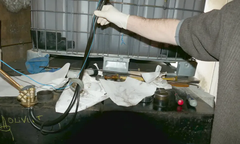 Main d'une personne en gant blanc manipulant un tube avec des fils bleus, près d'une cuve à fioul et d'une crépine.