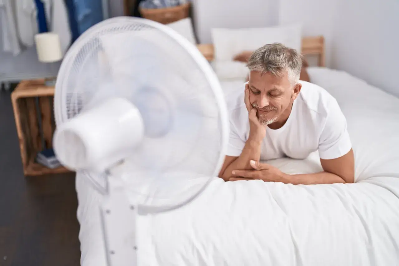 Un homme se repose sur un lit blanc, profitant de la brise d'un ventilateur. Une astuce simple pour comment rafraîchir une chambre quand il fait chaud.