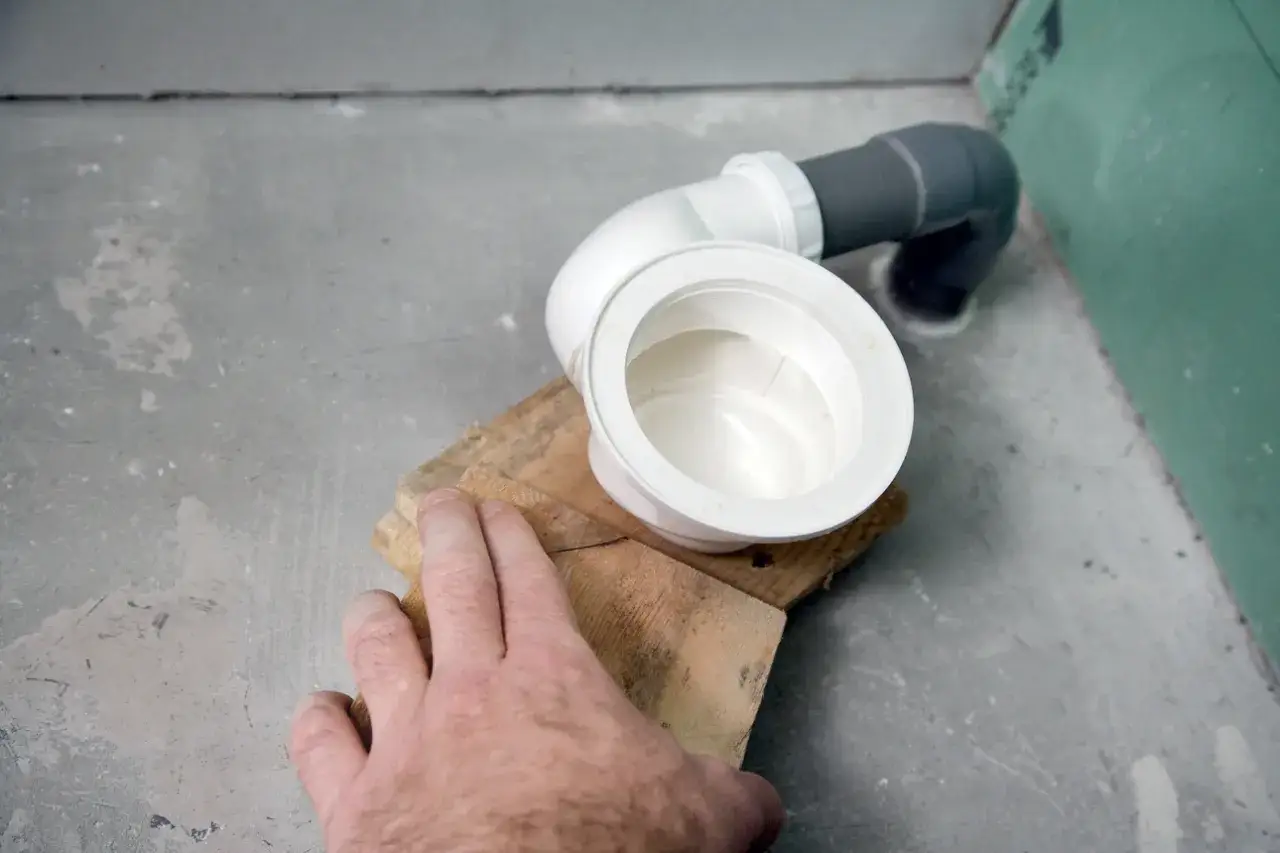 Main d'un artisan ajustant la pose du receveur douche, avec un siphon et des tuyaux en PVC.