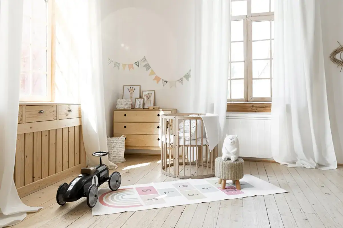 Chambre d'enfant lumineuse avec un radiateur sous fen&ecirc;tre en bois, un berceau rond, une commode, un tapis de jeu et une voiture jouet.