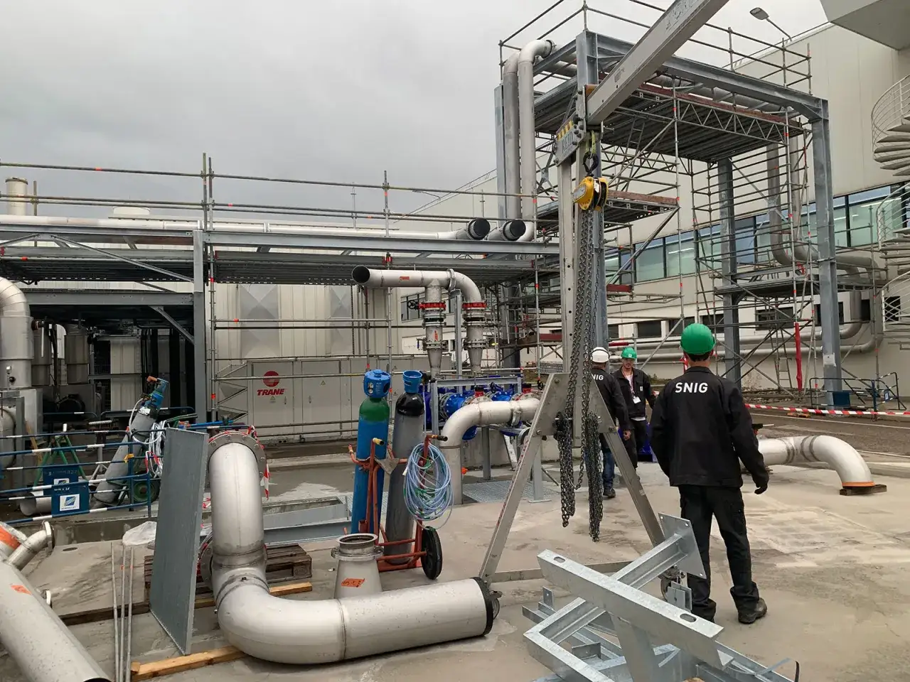 Chantiers industriels avec tuyauterie, échafaudages et personnel portant des casques. Un chariot élévateur est visible, ainsi que des bouteilles de gaz. Le logo SNIG est présent sur un vêtement.