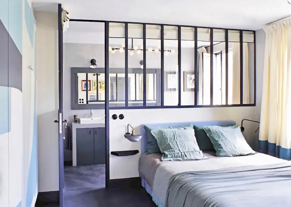 Chambre avec salle de bain ouverte sur chambre s&eacute;par&eacute;e par une verri&egrave;re. Lit bleu, coussins verts, rideaux clairs.