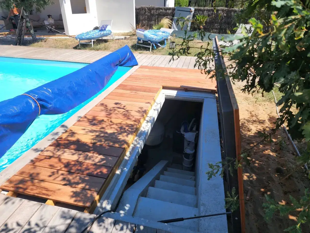Accès à un local technique de piscine, avec escalier en béton menant à l'intérieur. Une bâche bleue recouvre la piscine.