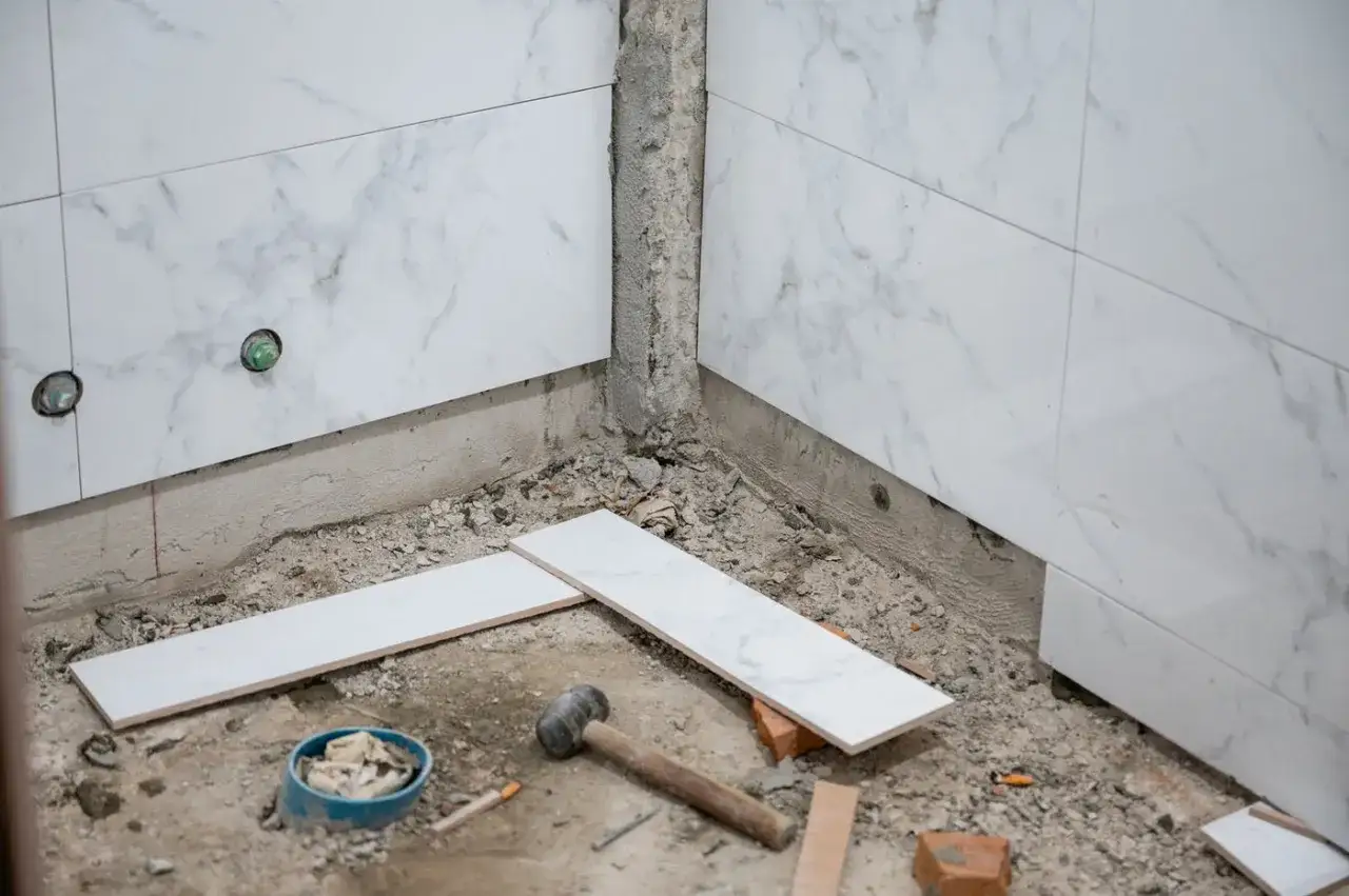 Des carreaux de marbre blanc sont prêts à être posés dans une salle de bain en construction. Un marteau et des outils sont au sol.