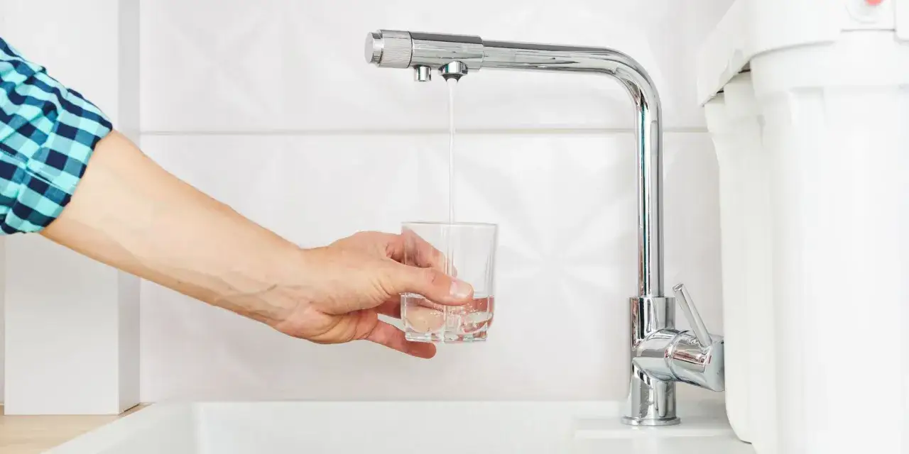 Main tenant remplissant un verre d'eau fraîche sous un mitigeur moderne, à côté d'un filtre à eau.