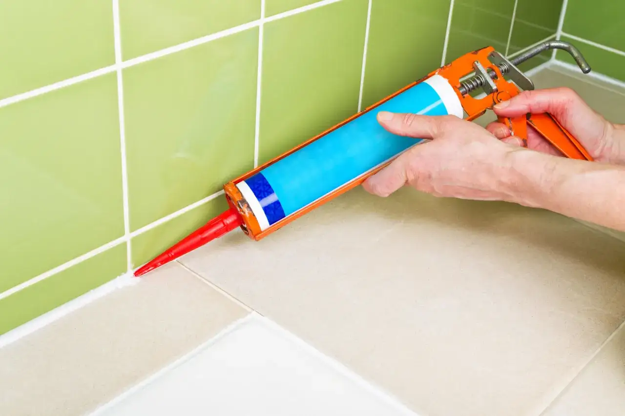 Main tenant utilise un pistolet &agrave; joint pour appliquer un cordon de mastic blanc entre des carreaux verts et beiges.