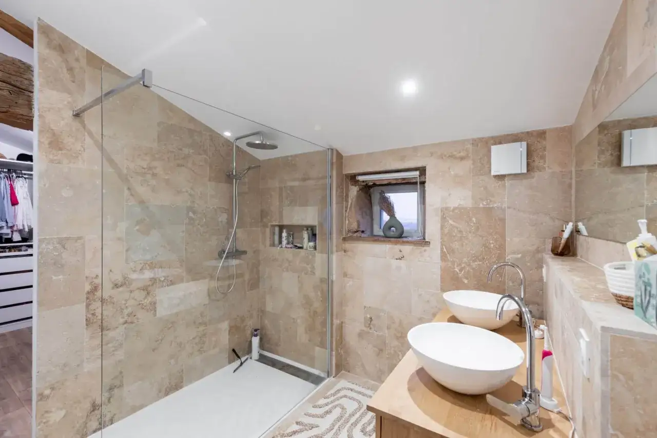 Salle de bain moderne avec douche vitrée, deux lavabos sur un meuble en bois, et murs en travertin. Idéal pour refaire salle de bain avec style.