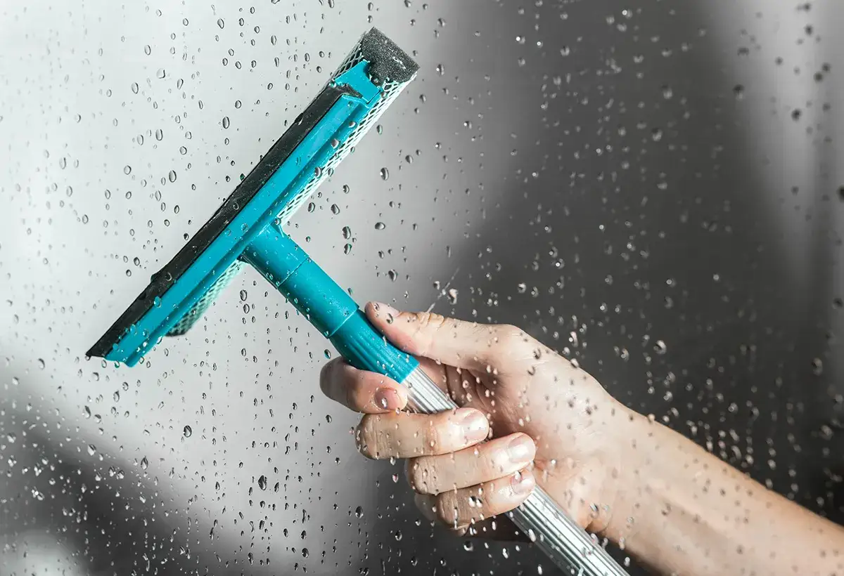 Main tenant une raclette pour enlever le calcaire de la douche sur une vitre mouillée.