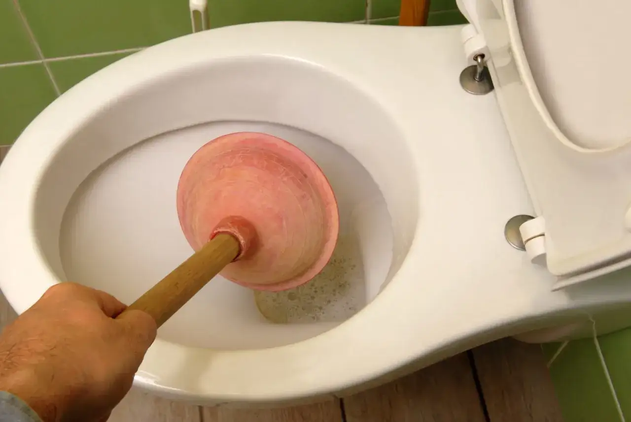 Main tenant, désespéré, tente de déboucher les toilettes avec une ventouse. L'eau trouble et mousseuse suggère une tentative infructueuse.