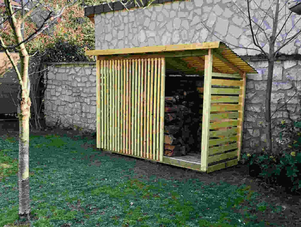 Abri bois de chauffage fait maison, avec façade à persiennes en bois, protégeant une belle réserve de bois sec.
