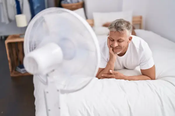 Un homme se repose sur un lit blanc, profitant de la brise d'un ventilateur. Une astuce simple pour comment rafraîchir une chambre quand il fait chaud.