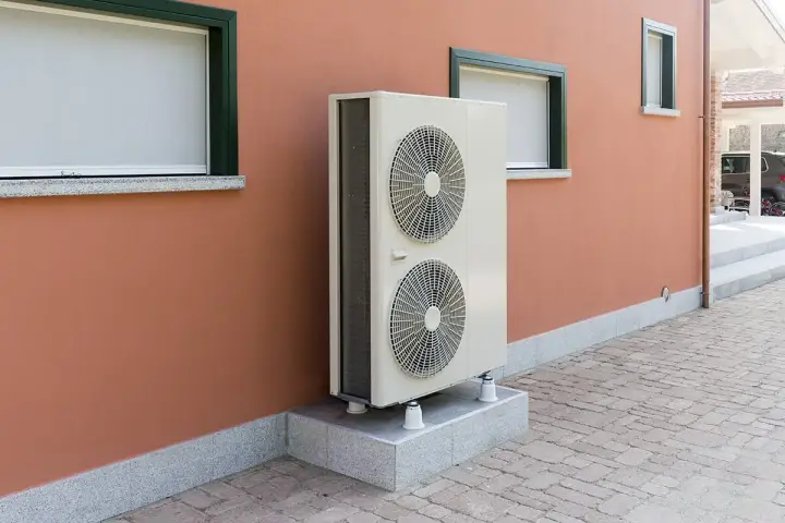 Unité extérieure de pompe à chaleur, avec ses deux ventilateurs, installée sur un socle devant un mur ocre. Le poids de cet appareil assure sa stabilité.