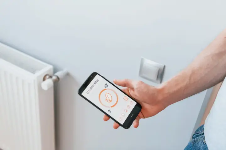 Main d'un homme tenant un smartphone affichant l'application Netatmo pour contrôler un radiateur électrique en mode économie.